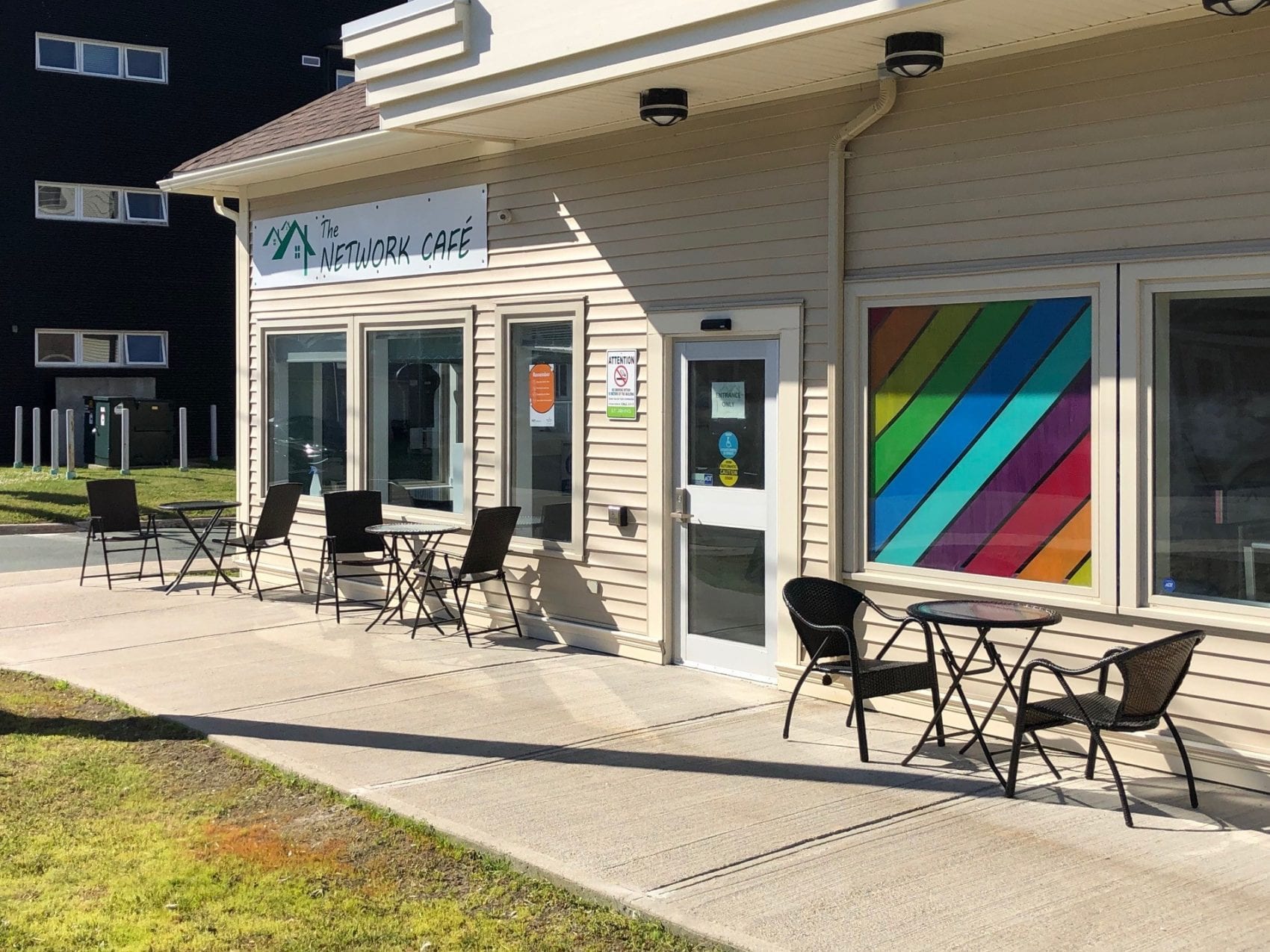 Am image of the patio of the Network Café. There are patio chairs and tables outside the entrance. A window is decorated in rainbow colors, and a sign reads "The Network Café" over the windows of the café.